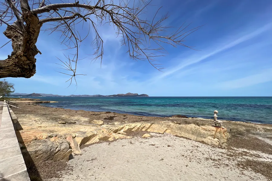 Felsplatten und heller Sand am Ufer im Frühjahr, links eine kahle Tamariske und die Promenadenmauer, rechts auf einem Felsvorsprung eine kleine sitzende Steinfigur, dahinter türkis und tiefblau abgestuftes Wasser.
