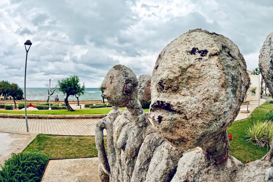 Grob behauene, hell verwitterte Steinfiguren mit runden Köpfen als Gruppe auf einer Grünfläche an der Promenade, dahinter Tamarisken, die Uferpromenade und das aufgewühlte Meer unter Wolkenhimmel.
