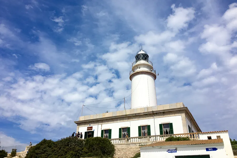 Der weiße Leuchtturm am Cap de Formentor von unten fotografiert, darüber ein blauer Himmel mit Schleierwolken, im Vordergrund das Turmgebäude mit grünen Fensterläden.
