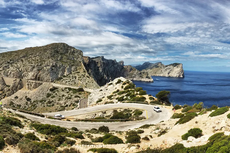 Weite Kehre der Straße zum Cap de Formentor mit Parkbucht, dahinter die zerklüftete Felsküste der Halbinsel und das offene Meer.
