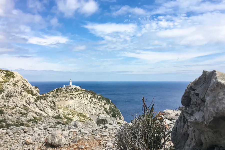 Blick über zerklüftete helle Felsen hinweg auf den fernen weißen Leuchtturm am Cap de Formentor, dahinter das offene Meer unter Wolkenhimmel.
