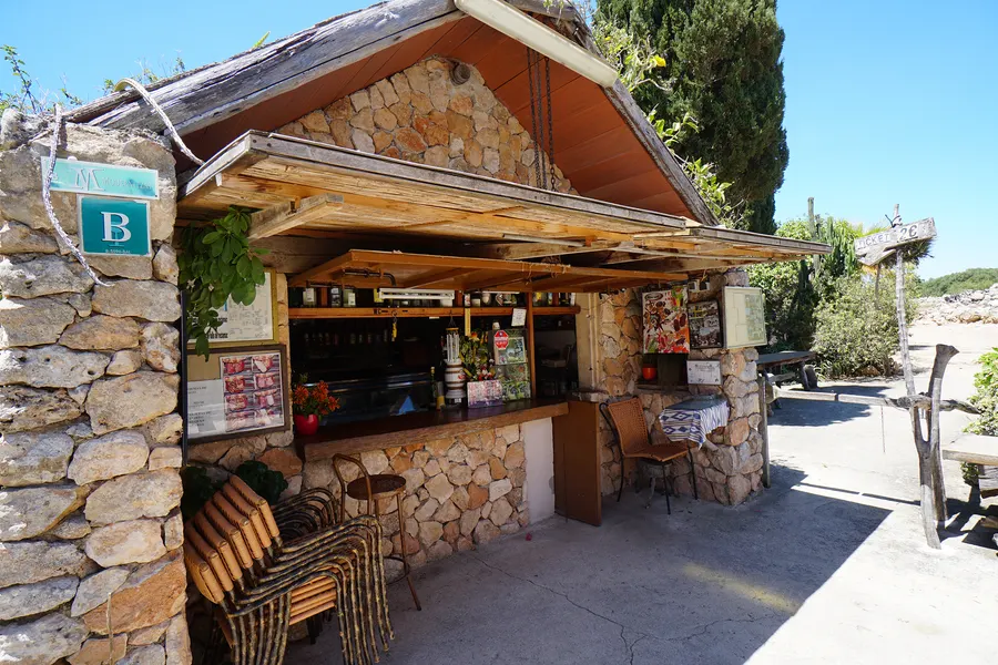 Kleine Bar aus Naturstein mit Holzdach am Eingang von Capocorb Vell, davor Tische und ein Hinweisschild.
