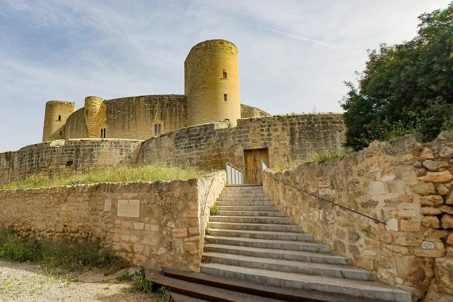 Steinerne Treppe, die zum Tor des Castell de Bellver hinaufführt, links die Burgmauer, rechts eine Trockenmauer und Bäume.