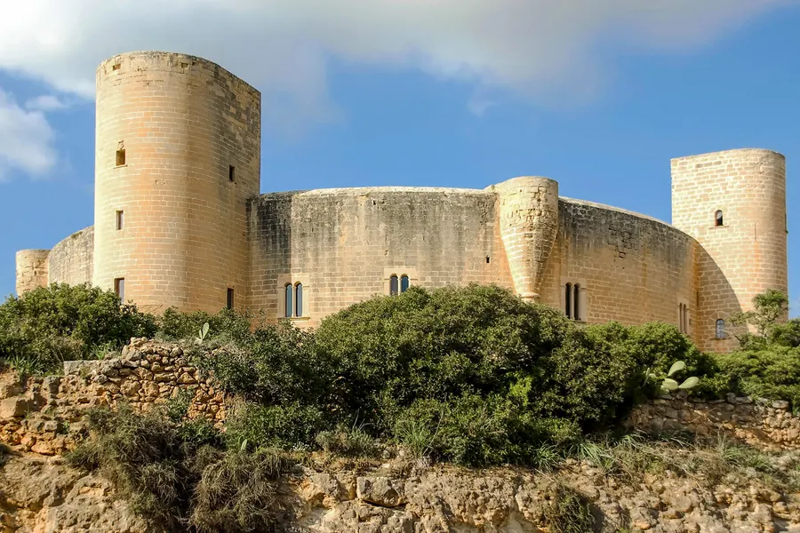 Das Castell de Bellver von unten gesehen über einem bewachsenen Felshang, Türme und Mauerring vor blauem Himmel.
