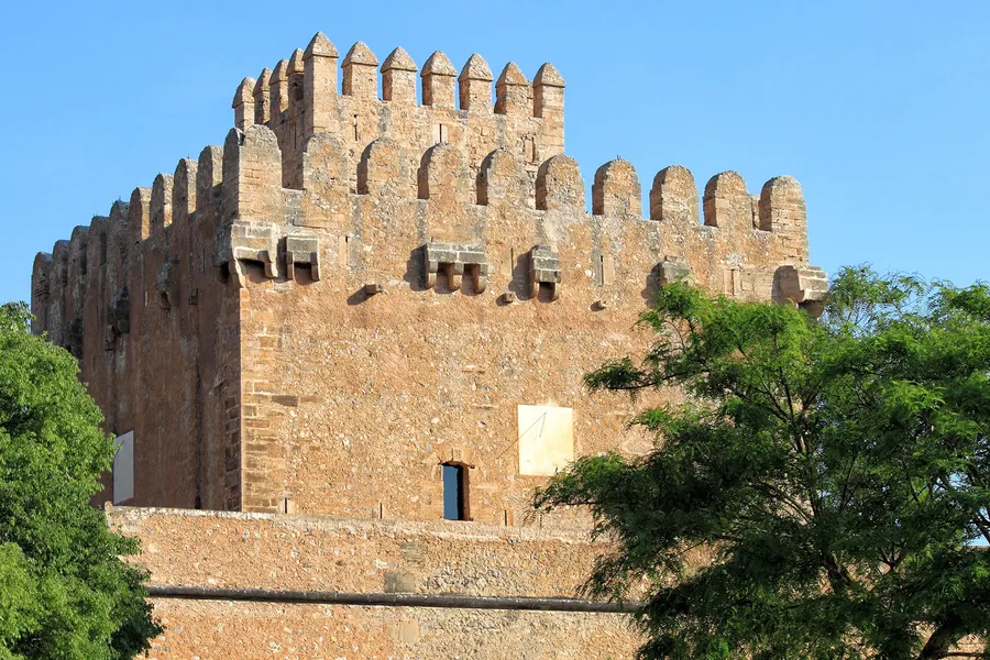 Der wuchtige quadratische Wehrturm Miquel Nunis aus hellem Sandstein mit Zinnenkranz und vorkragenden Konsolen, teilweise von Bäumen verdeckt.