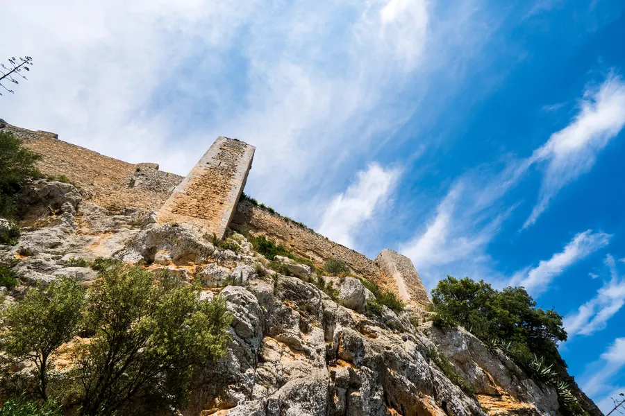 Ein hoher Mauerturm des Castell de Santueri über dem Felsabbruch, davor Buschwerk und trockenes Gras.