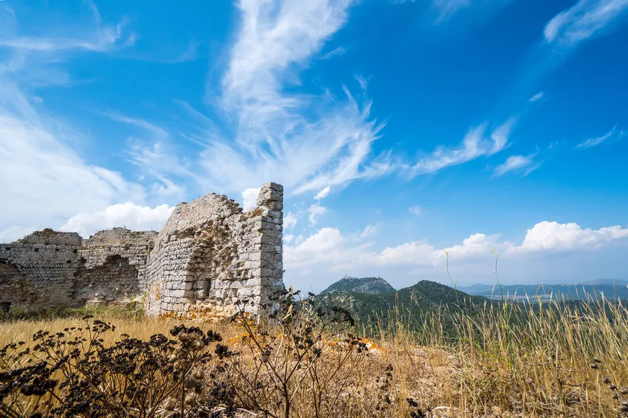 Freistehender Mauerrest des Castell de Santueri über trockenem Gras, dahinter Hügel und die Ebene im Dunst.
