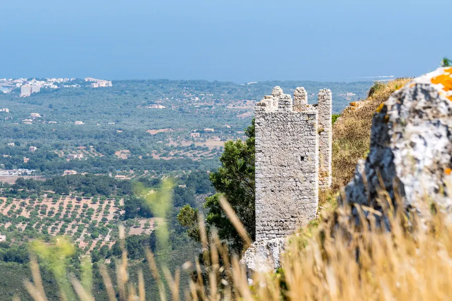 Zinnenbewehrter Mauerturm des Castell de Santueri hinter trockenen Gräsern, dahinter die Ebene mit Ortschaften.
