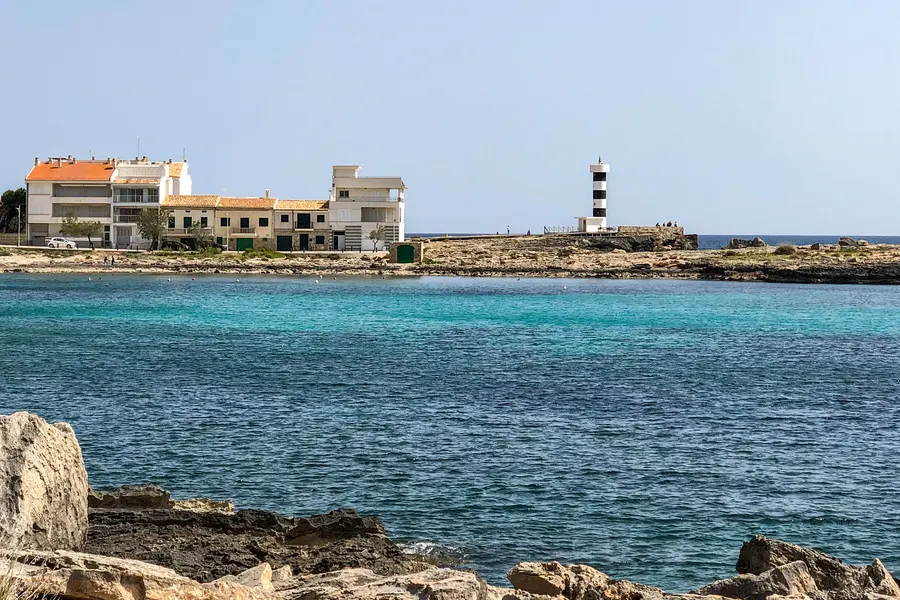 Blick über flaches klares Wasser auf die Häuser von Colònia de Sant Jordi und den gestreiften kleinen Leuchtturm an der Hafeneinfahrt.