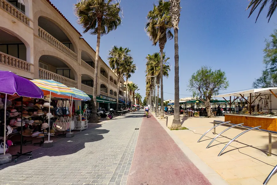 Sandstrand bei Colònia de Sant Jordi mit Holzsteg, Liegen und einem großen weißen Hotelbau unmittelbar dahinter.
