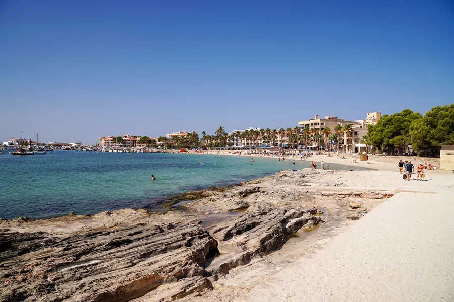 Der Ortsstrand von Colònia de Sant Jordi mit hellem Sand und flachen Felsplatten im Vordergrund, dahinter die Häuserzeile mit Palmen und der Hafen mit Booten.
