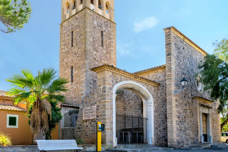 Bruchsteinkirche mit hohem Glockenturm, weiß gefasstem Rundbogenportal und Ziegeldach, davor eine Fächerpalme, eine weiße Bank und eine Gedenktafel zum ersten Jahrhundert des Orts.