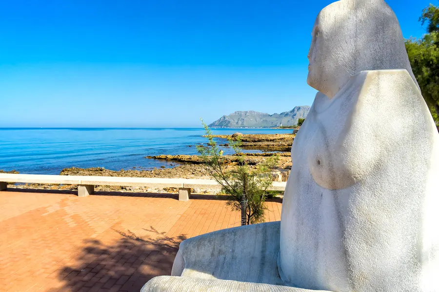 Große weiße Steinfigur einer sitzenden Frau auf der rot gepflasterten Uferpromenade, im Blick über die Felsküste hinaus auf das ruhige Meer und die Berge der Halbinsel Victòria.
