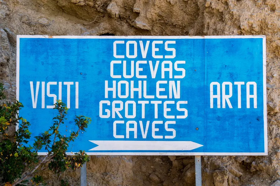 Verwittertes blaues Hinweisschild mit der Aufschrift Coves, Cuevas, Höhlen, Grottes, Caves und einem Pfeil Richtung Artà.