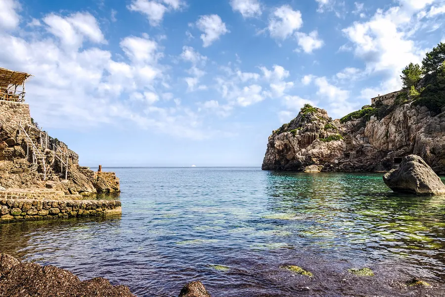 Die Cala de Deià mit klarem grünem Wasser zwischen hohen Felswänden, am Ufer ein Steg und einfache Bauten.