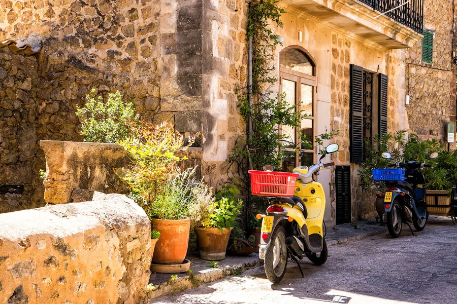 Enge Gasse in Deià mit Natursteinhäusern, Kübelpflanzen und abgestellten Motorrollern am Straßenrand.
