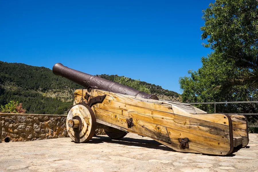 Alte Kanone auf einer hölzernen Lafette an einem Aussichtspunkt in Deià, dahinter bewaldete Hänge und Himmel.
