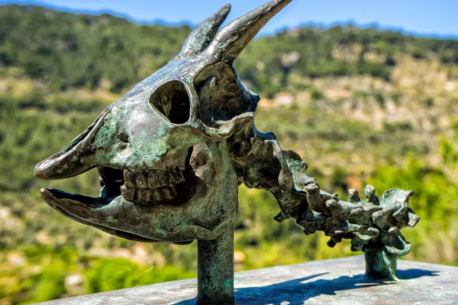 Bronzene Skulptur eines stilisierten Tierschädels auf einer Brüstung in Deià, dahinter unscharf das bewaldete Tal.
