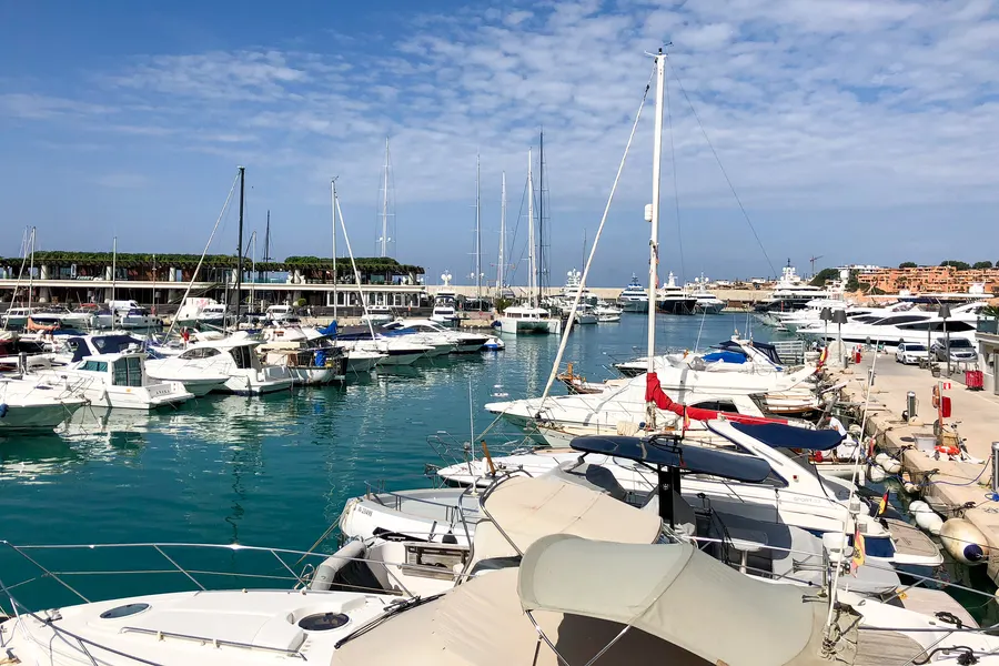 Gepflasterte Hafenpromenade in Port Adriano mit Läden, Lokalen und Booten auf der anderen Seite.
