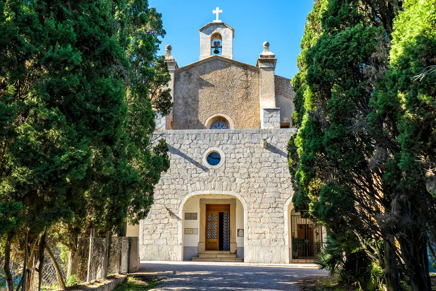 Frontalansicht der Ermita de Betlem: schlichte Bruchsteinfassade mit Rundbogeneingang, kleinem Rundfenster und Glockengiebel, links und rechts Zypressen und Zaun.