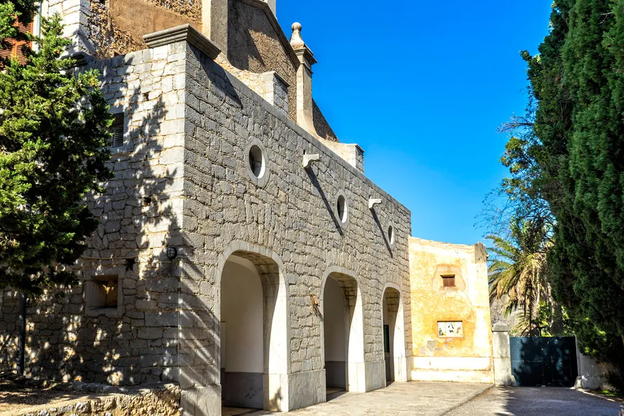 Seitenansicht der Klosteranlage von Betlem mit einer Reihe offener Rundbogenarkaden aus Naturstein, dahinter ein ockerfarbenes Nebengebäude.
