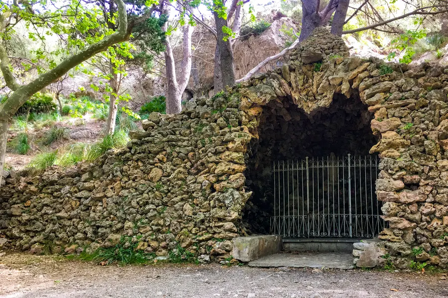 In eine Trockensteinmauer eingelassene Lourdesgrotte mit schmiedeeisernem Gitter, davor ein gepflasterter Weg unter Bäumen.
