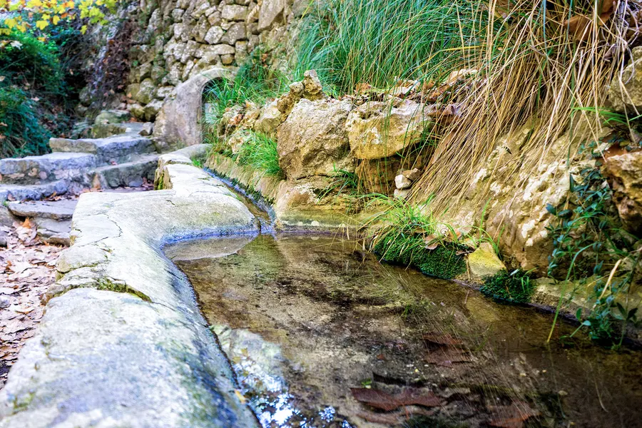 Steinerne Rinne mit klarem fließendem Wasser neben einem Felsen, umgeben von Gräsern und Moos.
