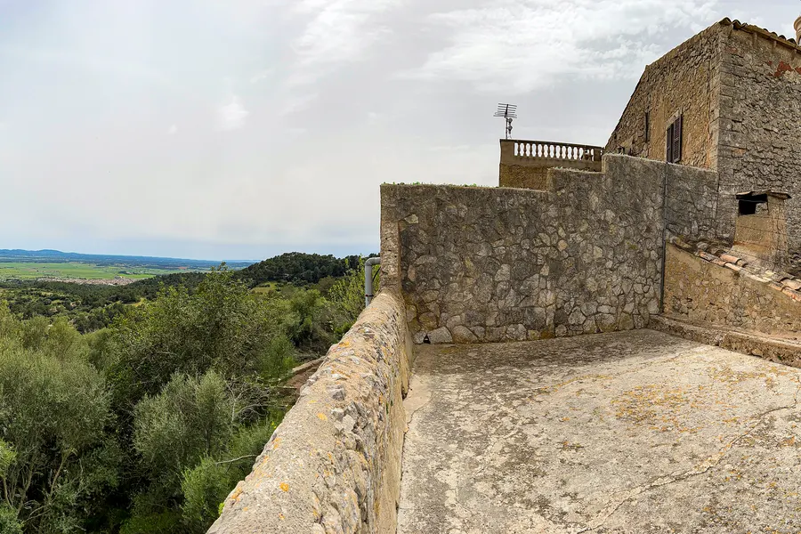Steinterrasse und Bruchsteinmauer der Ermita de Bonany, links fällt der Hang bewaldet ab, im Hintergrund die weite Ebene.