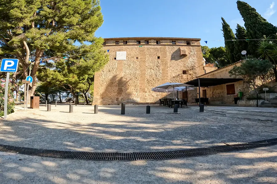 Blick vom Parkplatz auf die Rückseite der Anlage: massiver Bruchsteinbau mit Sonnenuhr und Fensterreihe unter dem Dach, davor eine Terrasse mit Sonnenschirmen und Tischen, links Kiefern und ein Parkplatzschild.