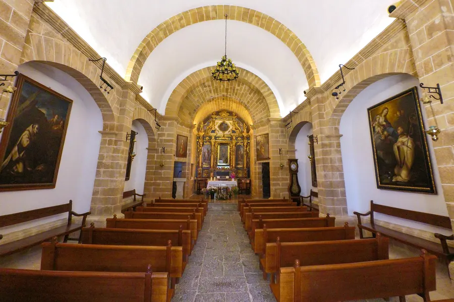 Schlichtes einschiffiges Kircheninneres mit Tonnengewölbe aus hellem Sandstein, Holzbänken, schmiedeeisernem Leuchter und einem vergoldeten Barockaltar am Ende; an den Wänden große dunkle Gemälde.