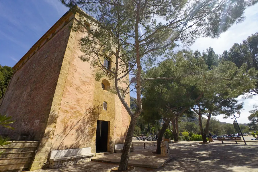 Hohe, fensterlose Seitenwand der Kirche aus rötlichem Bruchstein mit Sandsteinkanten und schmalem Portal, davor eine schräg gewachsene Kiefer; im Hintergrund der Vorplatz mit geparkten Motorrädern.