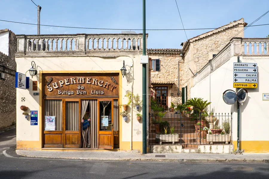 Kleiner Supermercat in Es Capdellà mit hölzernem Schaufenster und gemalter Aufschrift, daneben Wegweiser nach Andratx, Peguera, Calvià und Palma.