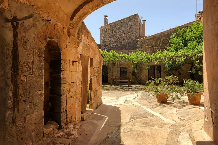 Gepflasterter Innenhof in Es Llombards mit Rundbogengang, Pflanzkübeln und einer Bank im Schatten unter Weinlaub.
