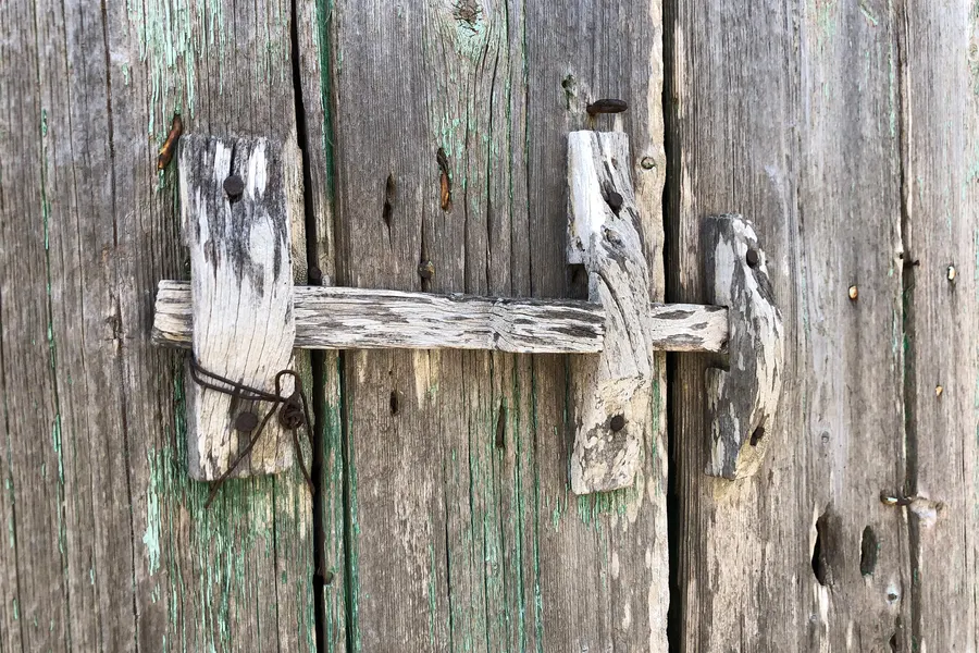 Nahaufnahme eines stark verwitterten Holztors mit grauen Brettern, grünen Farbresten und einem hölzernen Querriegel.
