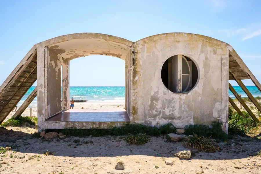 Betonskelett eines nie fertiggestellten Gebäudes am Strand von Ses Covetes, durch dessen Bögen das Meer zu sehen ist.
