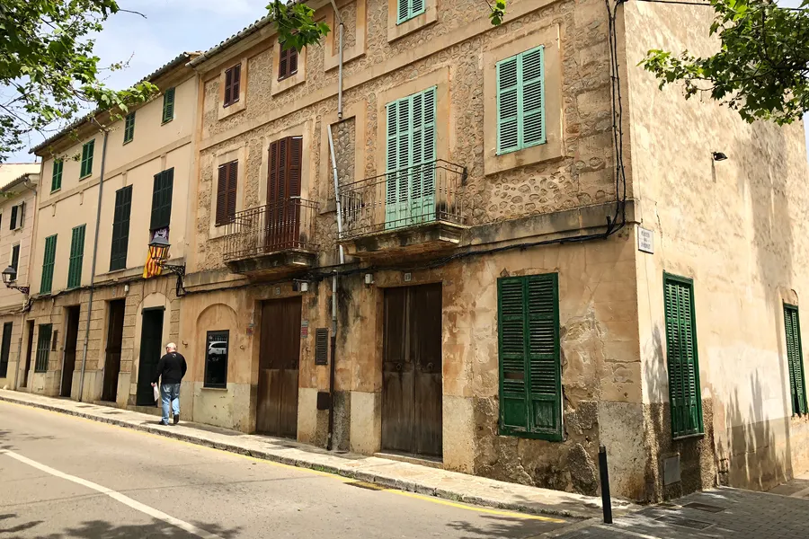 Straßenzug in Esporles mit hohen Häusern aus Naturstein, grünen Fensterläden, Balkonen und einem Passanten auf dem Gehweg.
