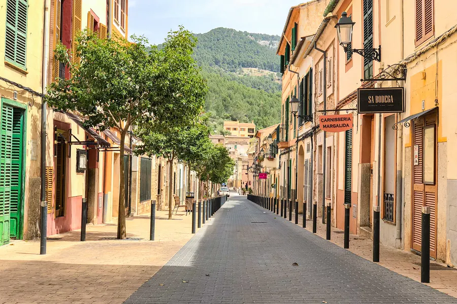 Gepflasterte Gasse in Esporles zwischen bunten Häusern, am Ende der Blick auf die bewaldeten Hänge der Serra de Tramuntana.