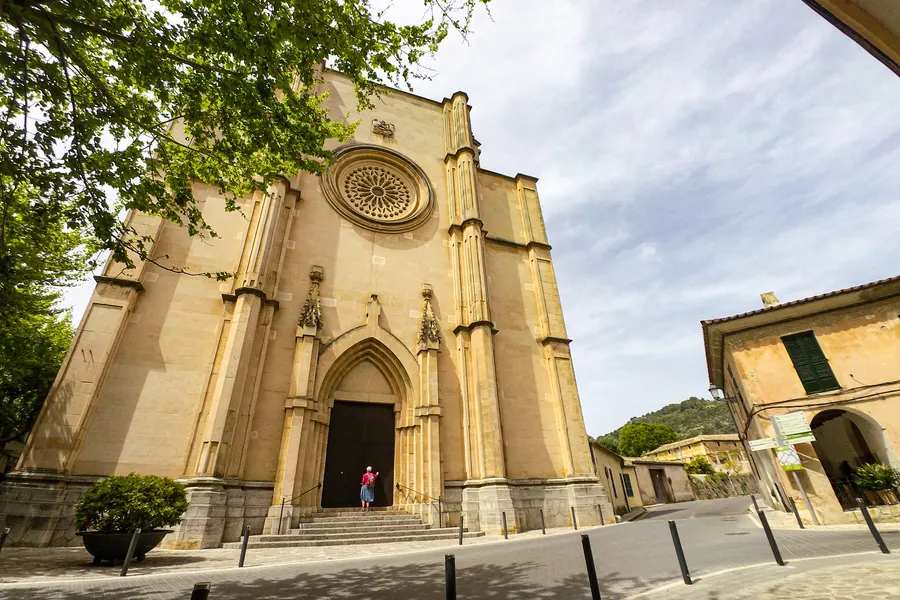 Die Pfarrkirche von Esporles aus hellem Sandstein mit spitzem Giebel, Rosettenfenster und Portal, davor Bäume und der Platz.
