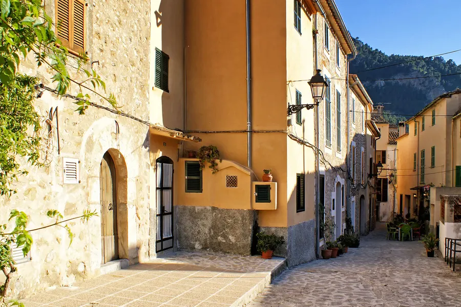 Ansteigende Gasse in Estellencs zwischen Natursteinhäusern, am Ende der Blick auf die bewaldeten Hänge der Serra de Tramuntana.
