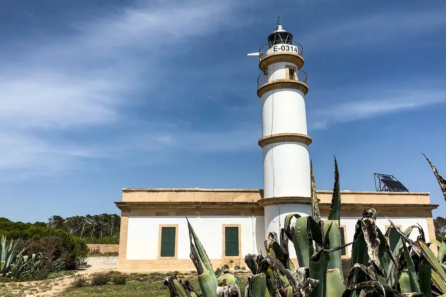 Der Leuchtturm am Cap de ses Salines mit dem angebauten Wärterhaus, grünen Fensterläden und Agaven im Vordergrund.