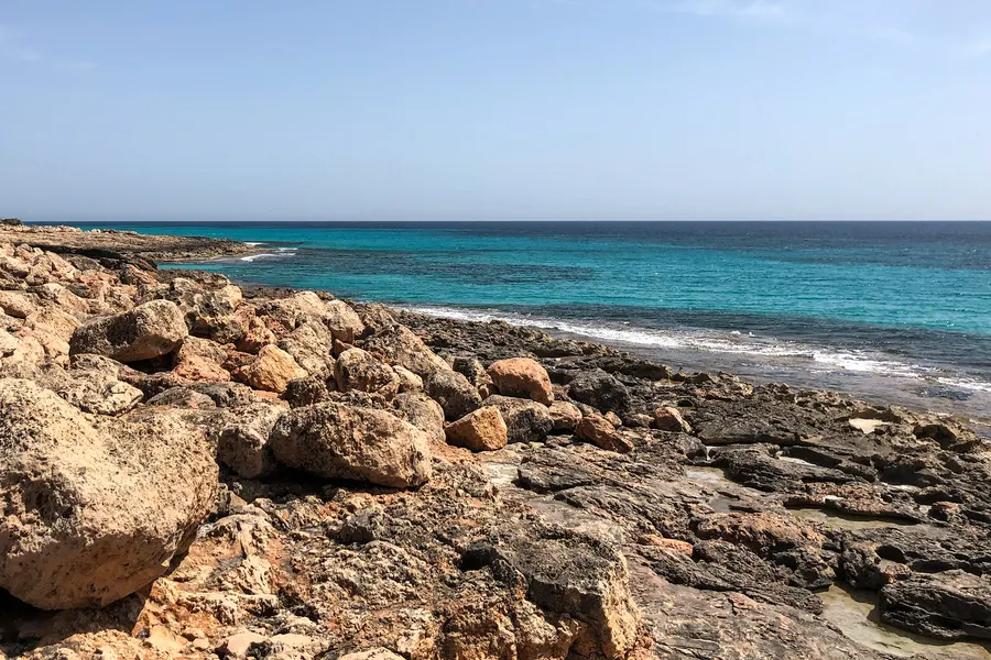 Zerfressene helle Felsplatten am Cap de ses Salines, dahinter türkisfarbenes flaches Wasser und ein heller Küstenstreifen.