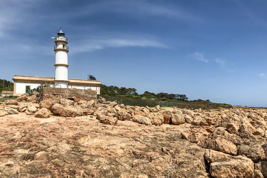 Der Leuchtturm des Cap de ses Salines von den vorgelagerten Felsplatten aus gesehen, unter weitem Himmel mit Schleierwolken.
