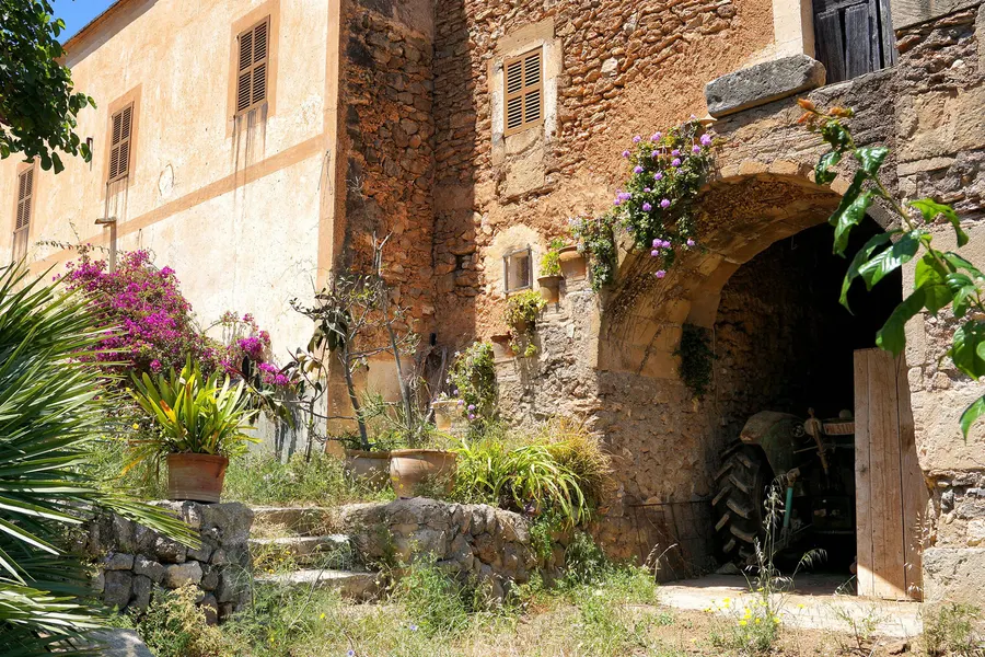 Hof in Felanitx mit hohem steinernem Torbogen, in dem ein Traktor steht, davor Bougainvillea und Topfpflanzen auf Trockenmauern.
