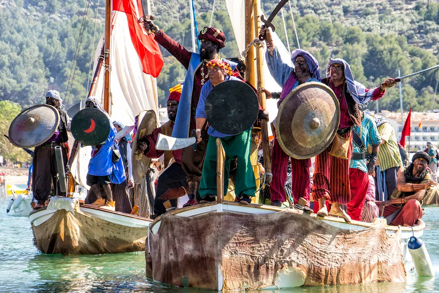 Zwei mit Fell bespannte Boote voller kostümierter Teilnehmer mit Rundschilden und Krummsäbeln in der Bucht von Port de Sóller beim Fest Es Firó.