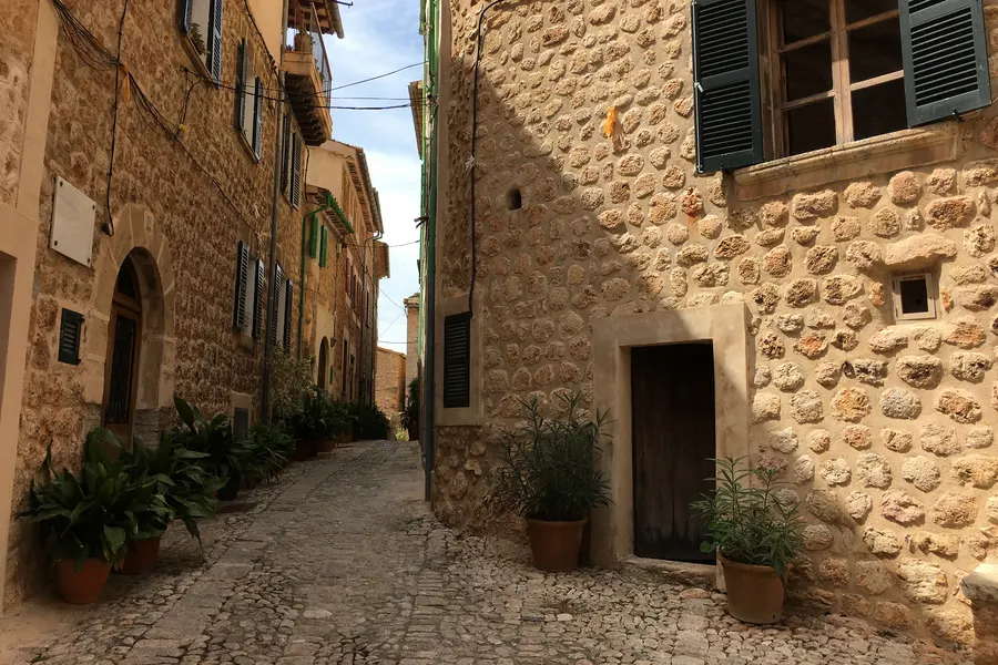 Gepflasterte Gasse in Fornalutx zwischen Bruchsteinhäusern mit dunklen Fensterläden und einer hölzernen Tür, davor Pflanzkübel.