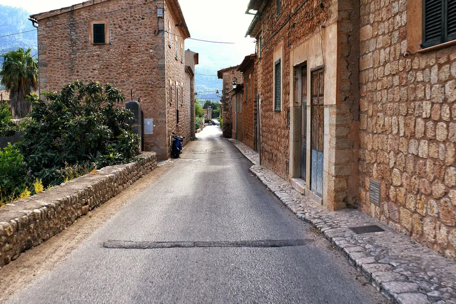 Straße durch Fornalutx zwischen Natursteinhäusern mit grünen Fensterläden, am Ende der Straße die bewaldeten Hänge der Tramuntana.
