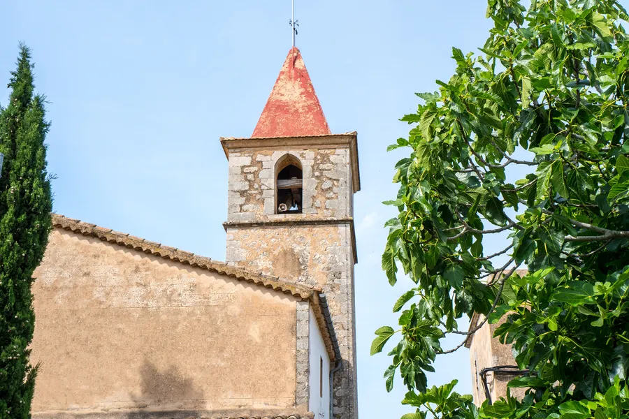 Der schlanke Glockenturm der Kirche von Galilea mit rotem Spitzdach und Wetterhahn, umgeben von Zypressen und Laub.