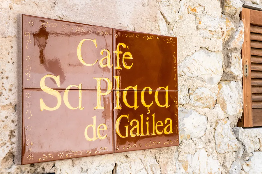 Handgemaltes Schild mit der Aufschrift Café Sa Plaça de Galilea an einer hellen Natursteinwand neben einem Fensterladen.