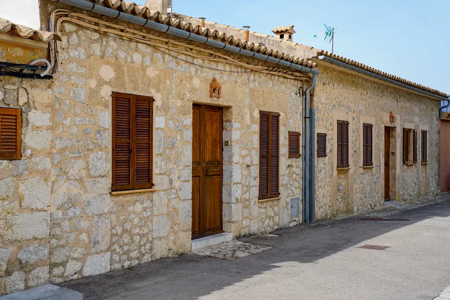 Langgestreckte Häuserzeile in Galilea aus hellem Naturstein mit hölzernen Fensterläden und Türen, davor die schmale Dorfstraße.
