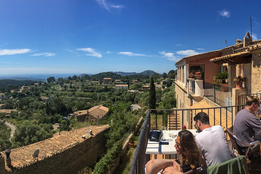 Gäste an Tischen auf einer Terrasse in Galilea, dahinter der freie Blick über Olivenhänge und die Ebene bis zum Horizont.
